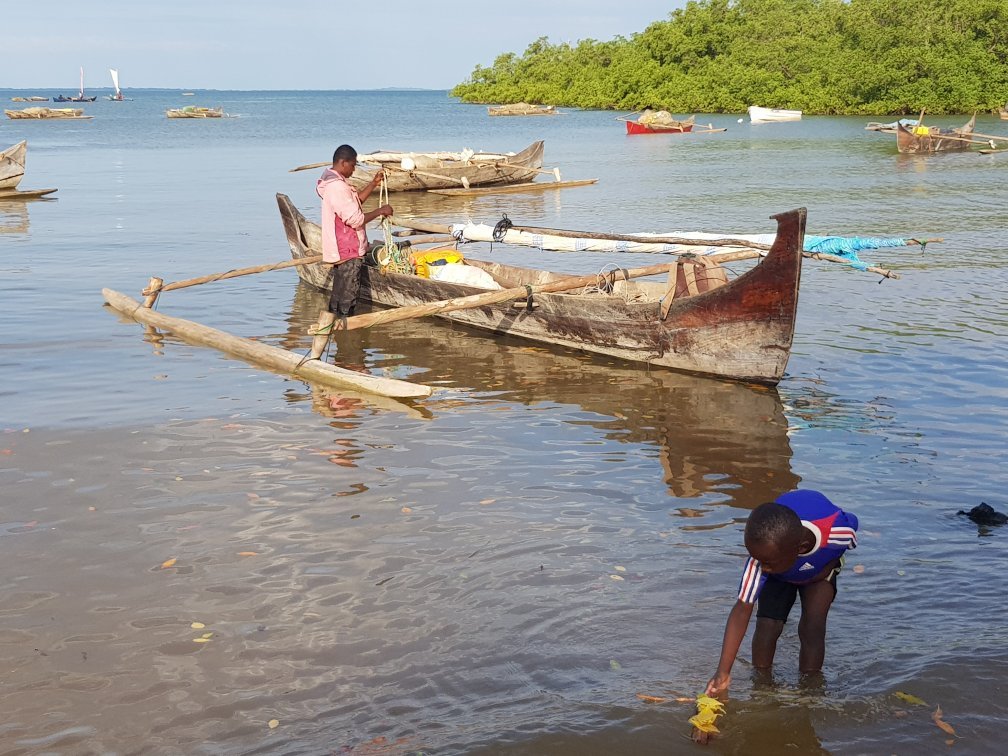 A Malagasy fisherman rigging the sail of his outrigger pirogue while a boy wades in the shallows beside him