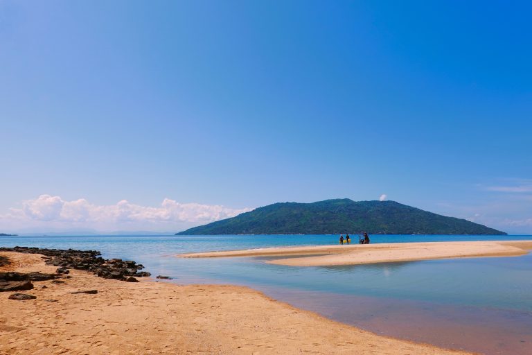 A curving golden sandspit reaching towards a forested island across a bay of clear turquoise water