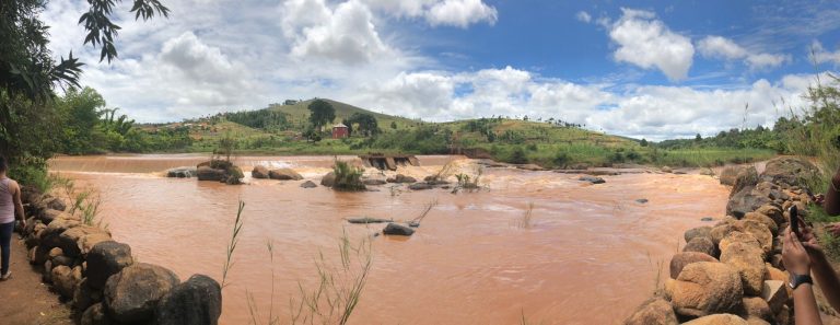 Red silt-laden water pouring over a low weir beneath a village on the hillside above