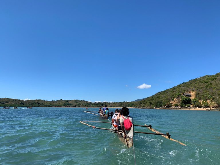Two loaded outrigger pirogues carrying visitors across a wide, sunlit bay