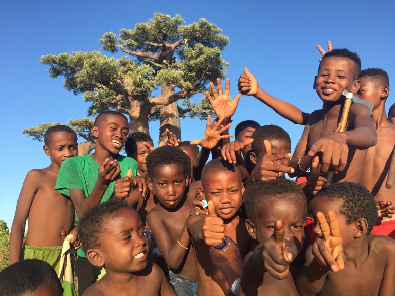 A group of village children laughing and cheering beneath a huge Baobab tree